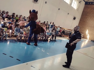 zdjęcie - sala gimnastyczna, dzieci, policyjna maskotka wita się z nimi, a policjant mówi przez mikrofon do dzieci