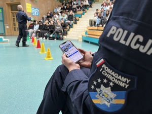 zdjęcie - policjantka z telefonem na którym wyświetlona jest strona śląskiej policji, w tle policjant opowiadający o zaletach pracy w policji