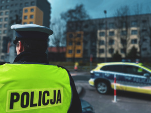 zdjęcie przedstawia policjanta drogówki stojącego przy szkole w rejonie przejścia dla pieszych, w tle widoczny radiowóz i budynki