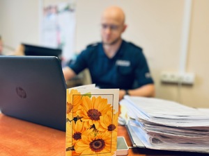 zdjęcie przedstawiające kartkę z podziękowaniami ustawioną na biurku policjanta, w tle policjant pracujący na komputerze, materiały kolejnych spraw